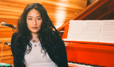 Chloe Flower seated in front of a red grand piano with sheet music, wearing a black jacket over a white top.